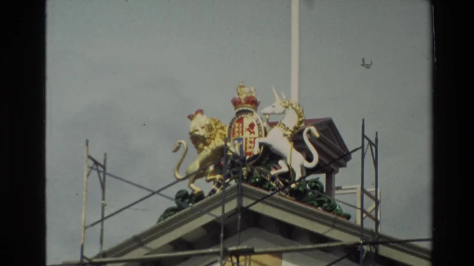 1981 New Zealand home-movie frame repeating the roofline coat of arms, with lion and unicorn figures, a central shield, scaffolding, and a flagpole.