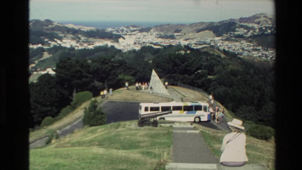 1981 New Zealand overlook scene with a white tour bus, visitors near a stone pyramid memorial, and a coastal city beyond hills.