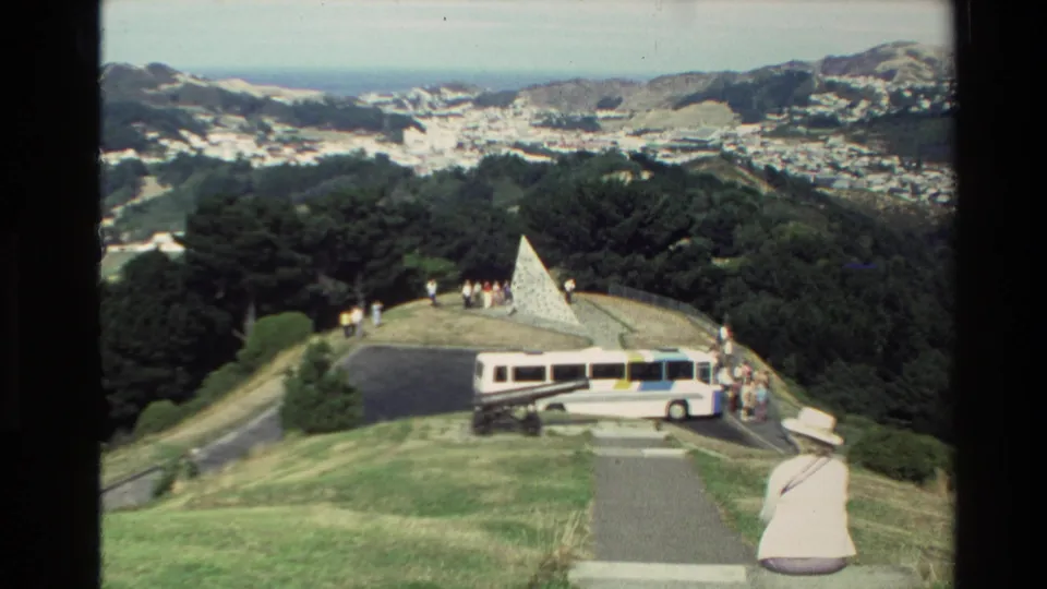 Wide 8mm frame of a New Zealand scenic viewpoint with parked tour bus, visitors, stone memorial, green hills, and distant city.