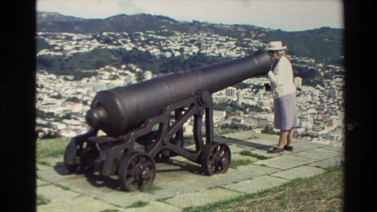 1981 New Zealand home-movie frame showing a woman in a white hat beside a large historic cannon overlooking a city and green hills.