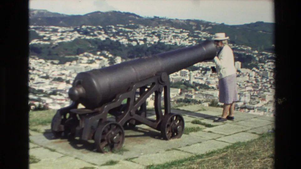 1981 New Zealand home-movie frame showing a woman in a white hat beside a large historic cannon overlooking a city and green hills.