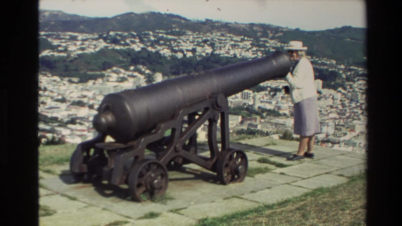 Woman standing beside a long cannon with a city and hills in the background.
