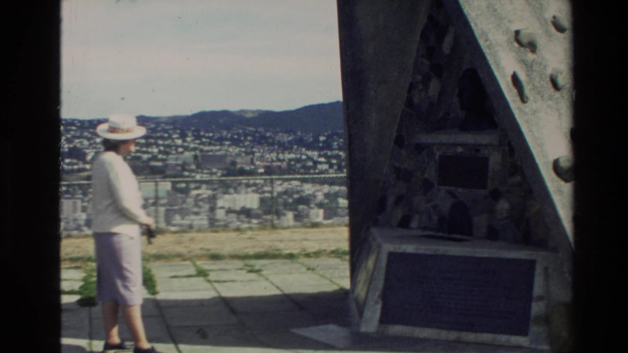 1981 New Zealand home-movie frame showing a woman in light clothing near a stone monument with a city panorama behind her.