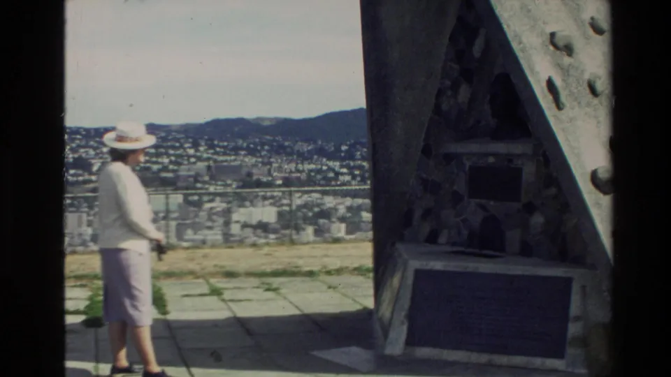 1981 New Zealand home-movie frame showing a woman in light clothing near a stone monument with a city panorama behind her.