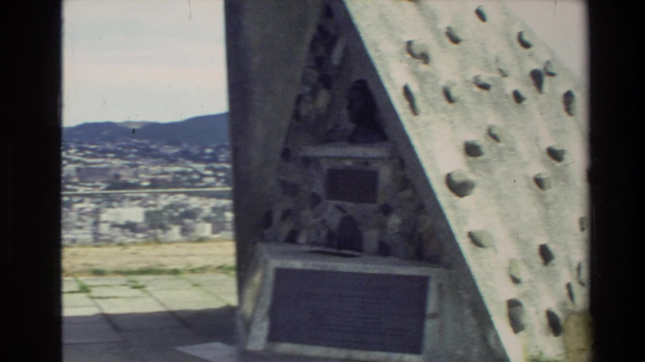 Triangular stone monument with inset plaques and a small bust appears at a New Zealand city overlook in 1981 home-movie footage.