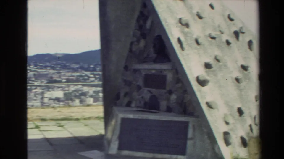 Triangular stone monument with inset plaques and a small bust appears at a New Zealand city overlook in 1981 home-movie footage.