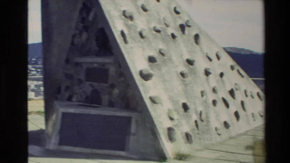 Close 8mm frame of a triangular stone monument with embedded stones, dark plaques, and paved viewing area in New Zealand.
