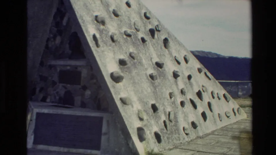 New Zealand home-movie frame from 1981 showing the monument's textured sloped side, plaques, and hills in the background.