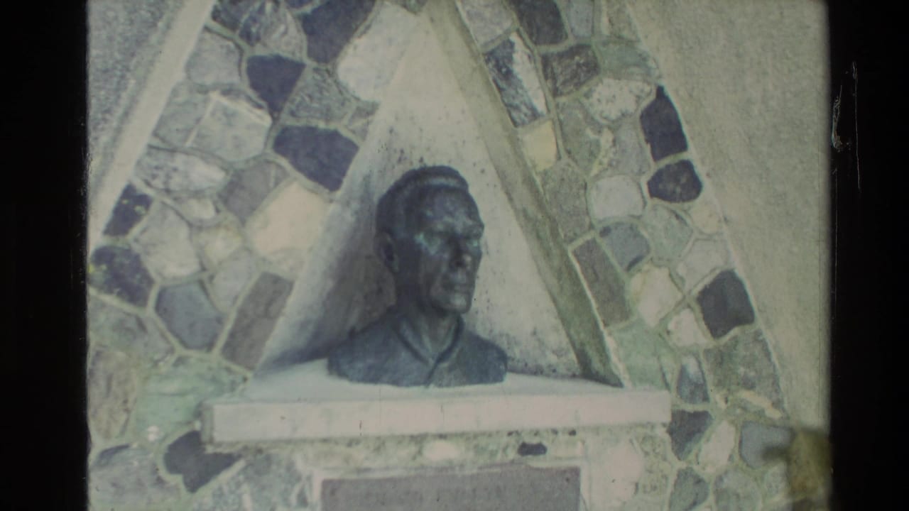 1981 New Zealand home-movie frame showing a sculpted bust in front of a patterned stone mosaic wall.