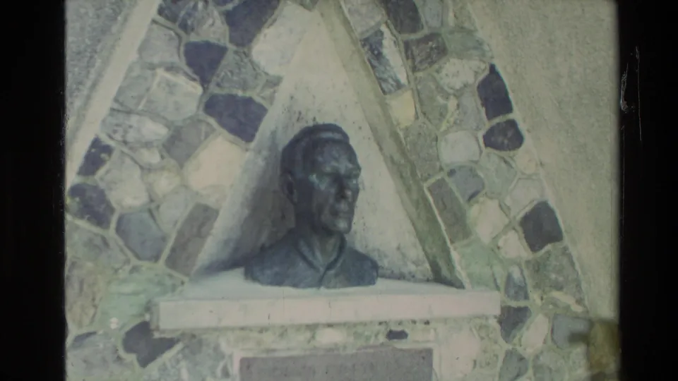 1981 New Zealand home-movie frame showing a sculpted bust in front of a patterned stone mosaic wall.