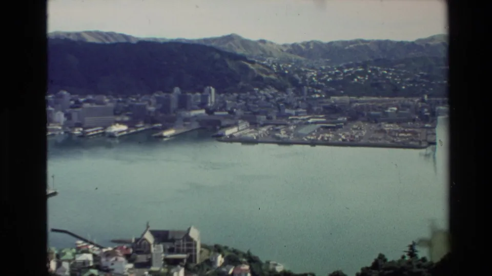 Elevated 1981 New Zealand view across a coastal city and bay, with piers, waterfront buildings, and hills beyond.