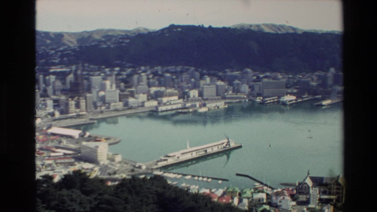 Coastal city skyline beside a harbor with a long ferry terminal and hills in the background.
