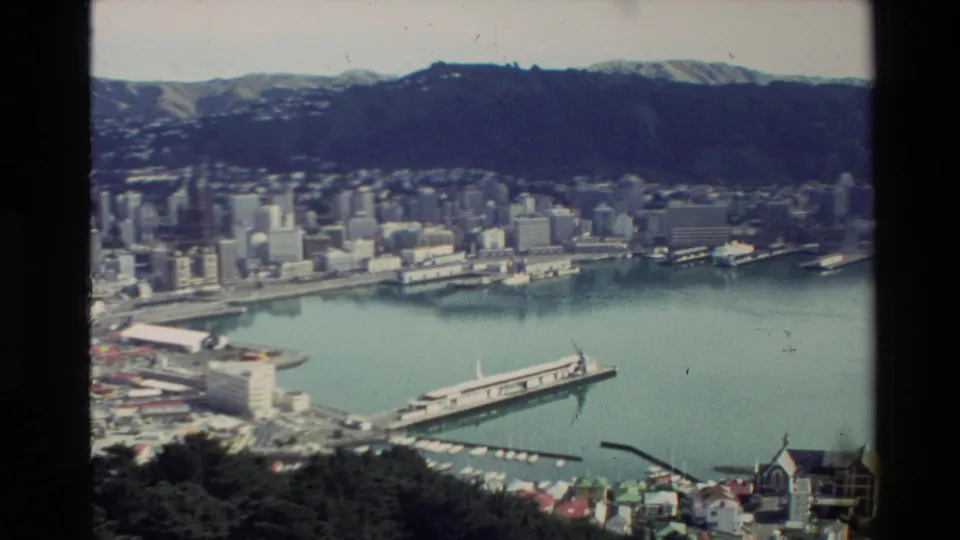 New Zealand harbor city seen from above in 1981, with a ferry wharf, mid-rise skyline, hillside homes, and mountain backdrop.