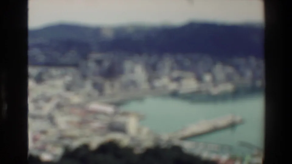Blurred 1981 8mm frame of a New Zealand harbor city from a high viewpoint, with waterfront, skyline, and hills softened by focus shift.
