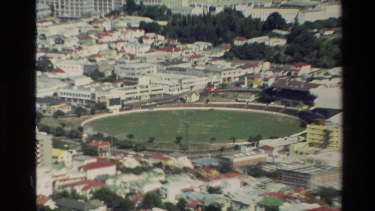 1981 New Zealand 8mm frame showing an oval stadium amid dense low-rise housing and city blocks from a high vantage point.