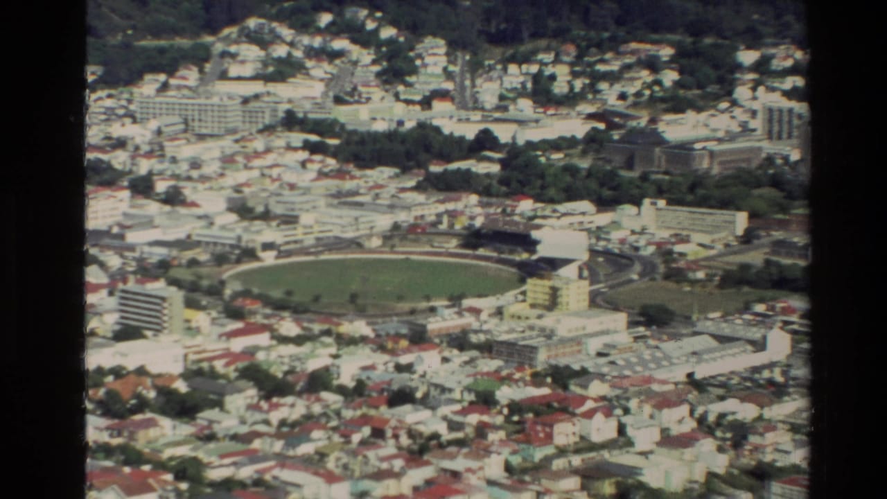 1981 New Zealand city panorama with a stadium in the middle distance, surrounded by houses, roads, and wooded hills.