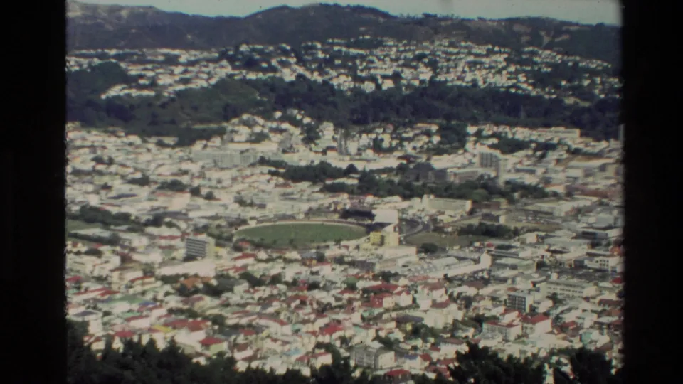 1981 New Zealand 8mm frame showing the stadium from farther back, with hillside housing and wooded slopes around the city.
