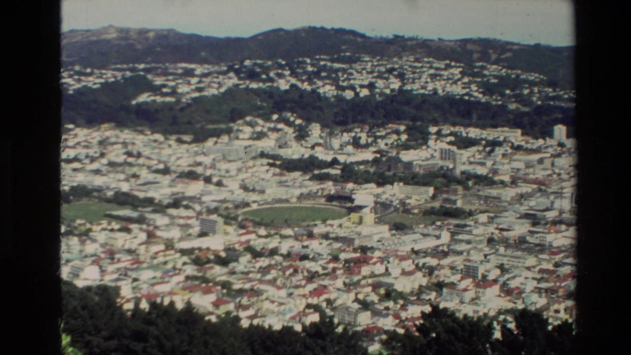 1981 New Zealand home-movie frame showing a city spread across hills, with rooftops, a sports ground, trees, and distant ridgelines.