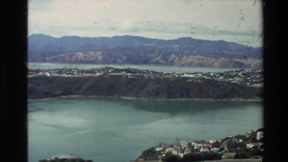 1981 New Zealand frame looking across a broad lake or harbor toward shoreline neighborhoods and mountain ridges.