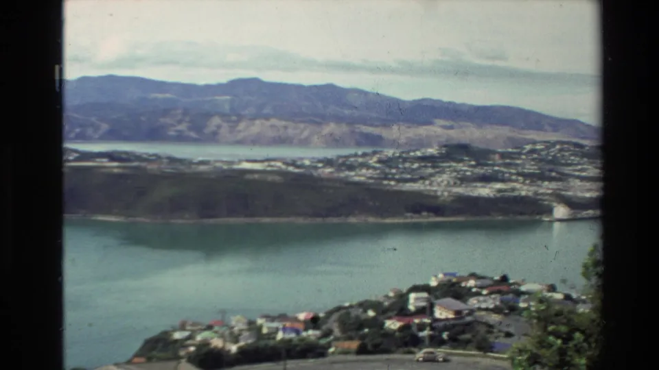 1981 New Zealand home-movie frame showing calm water, hillside suburbs, distant ridges, and foreground homes near a lookout.