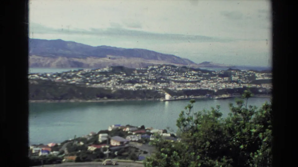 1981 New Zealand 8mm view of a coastal town beside a broad body of water, framed by dark film borders and foreground bush.