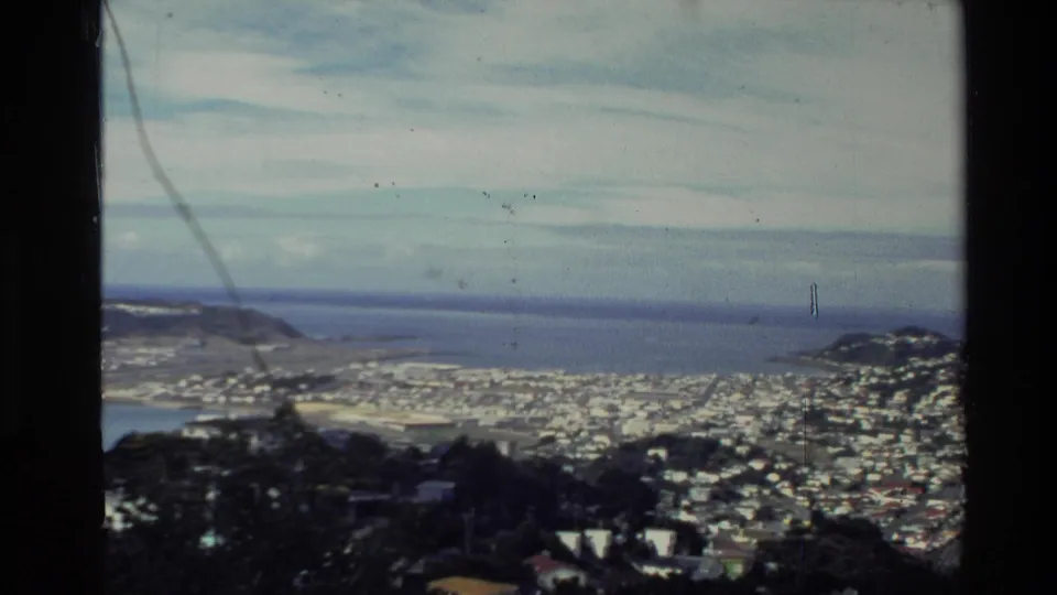 1981 New Zealand home-movie vista showing hillside homes, waterfront settlement, distant water, and visible 8mm surface marks.