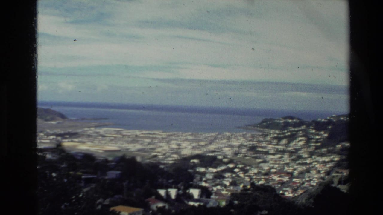 1981 New Zealand 8mm frame showing a coastal town spread across hills beside a broad bay under a cloudy sky.