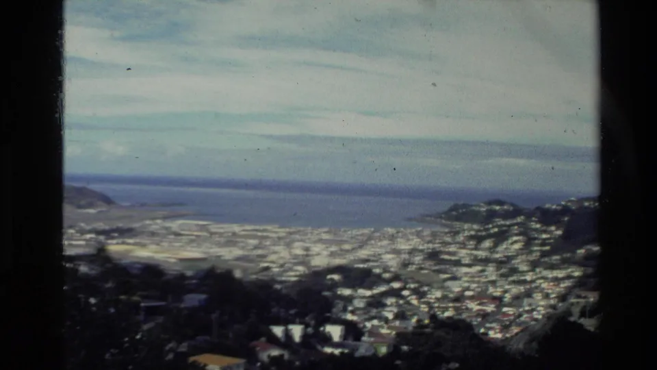 1981 New Zealand 8mm frame showing a coastal town spread across hills beside a broad bay under a cloudy sky.