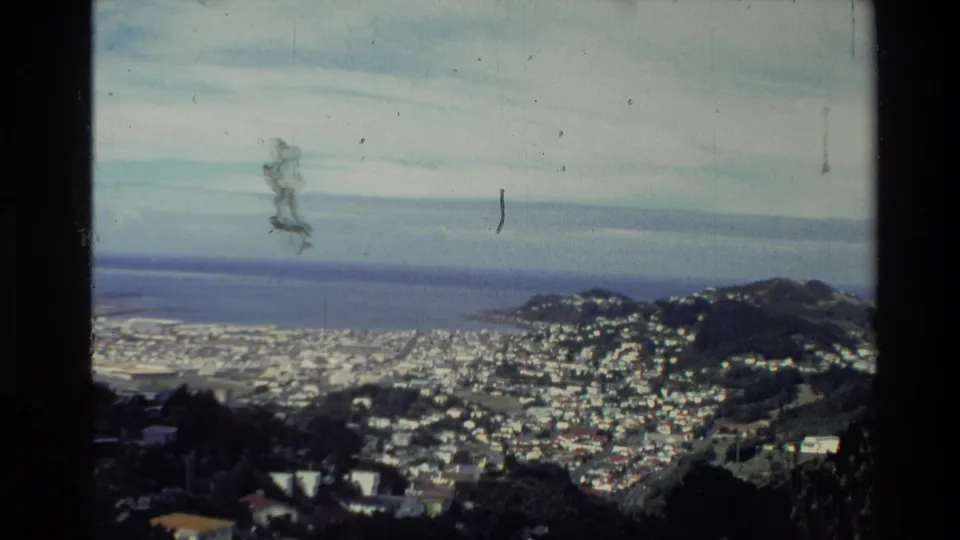 Elevated 1981 New Zealand view of a dense coastal settlement, bay water, small landforms, and visible film scratches.