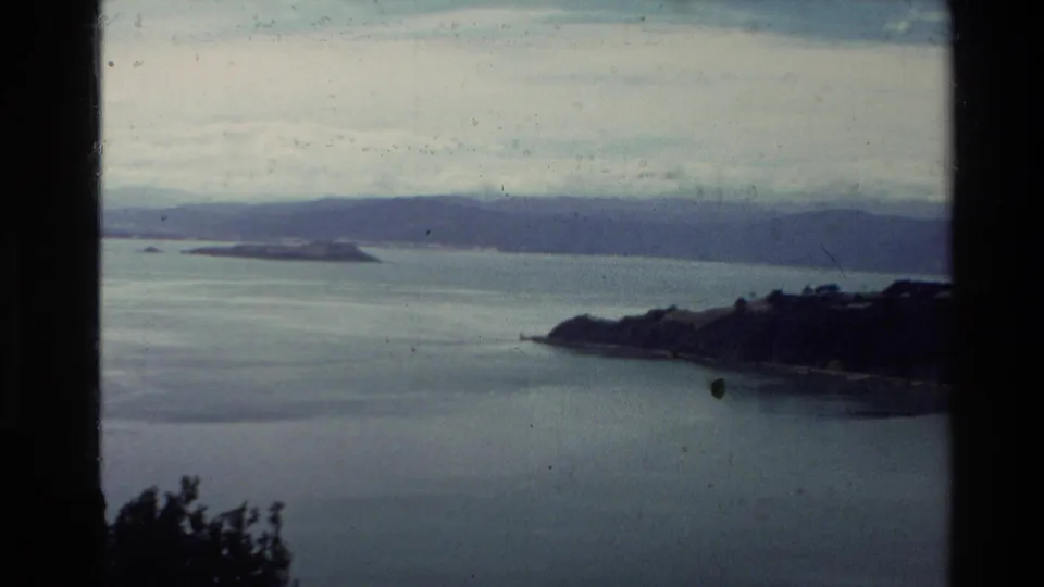 1981 New Zealand 8mm frame showing calm coastal water, distant hills, small islands, cloudy sky, and dark foreground framing.