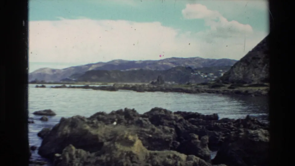 1981 New Zealand 8mm frame showing a rocky shore, calm water, distant hills, pale clouds, and a dark foreground edge.