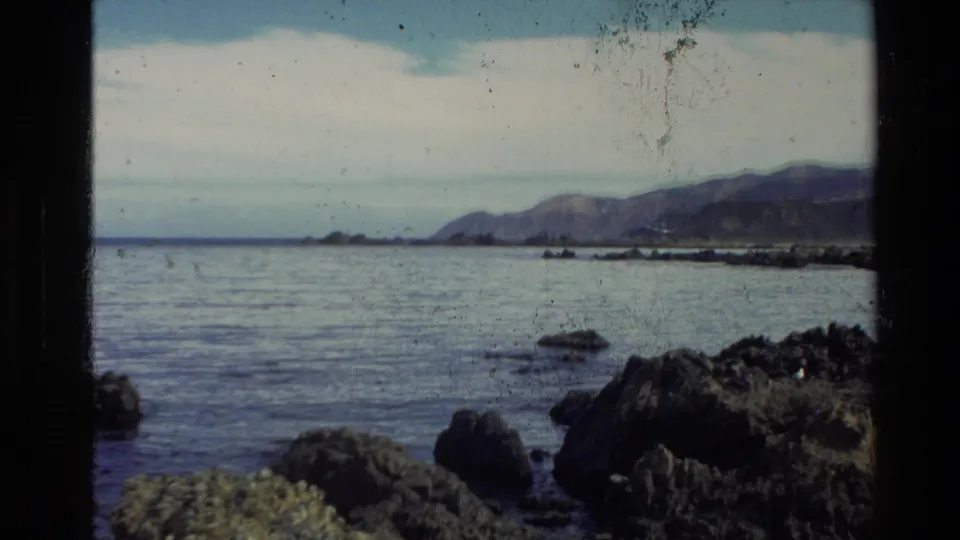 Archival 8mm view of a New Zealand shoreline, with rugged rocks in the foreground, calm sea, and mountains across the water.