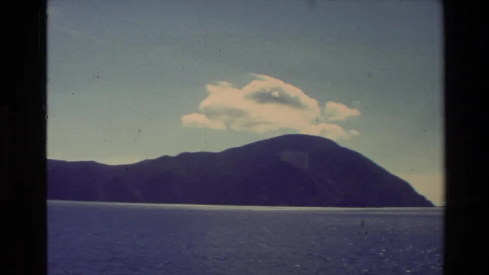A dark New Zealand mountain coastline rises beyond calm sea in 1982 8mm home-movie footage, with small clouds in a pale sky.