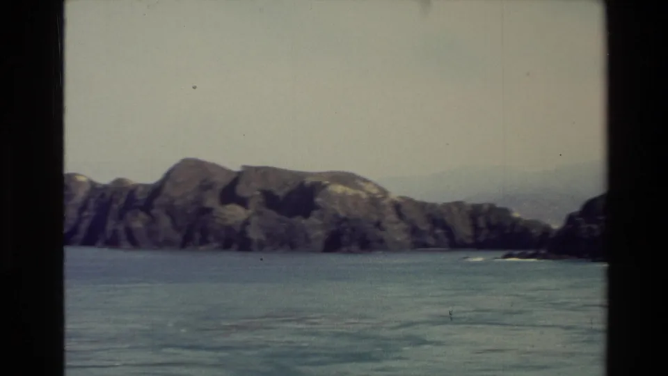 1982 New Zealand 8mm coastal view of rugged rocks and blue-green sea, framed by the moving boat and film gate.