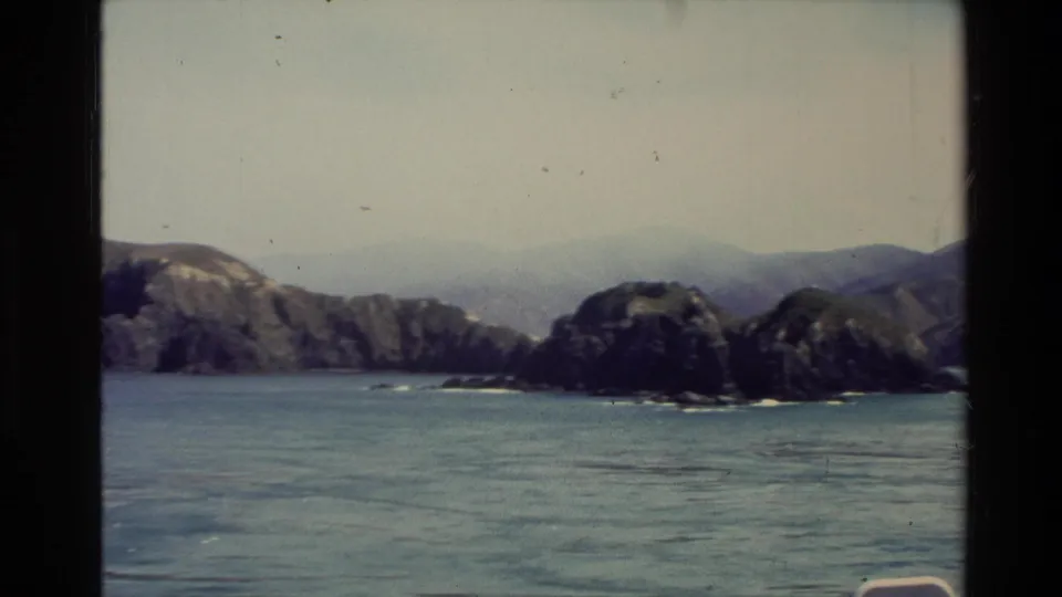 1982 New Zealand 8mm frame of rocky islands, turquoise water, and hazy inland hills from a boat viewpoint.