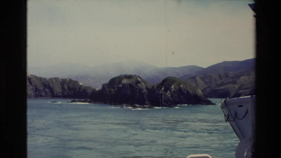 1982 New Zealand 8mm coastal frame showing a rocky island, distant mountains, open water, and a partial boat edge at right.