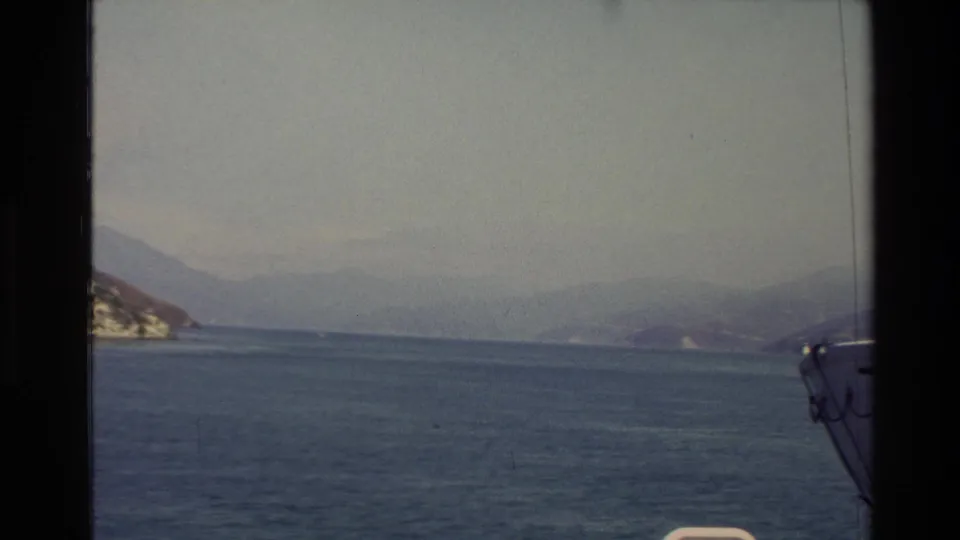 1982 New Zealand coastal view from a vessel, showing blue water, hazy mountains, rocky shoreline, and part of the boat at frame edge.