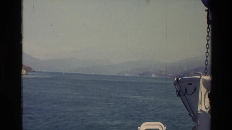New Zealand 1982 home-movie frame with calm blue water, distant mountains, hazy shoreline, and boat hardware visible at the right edge.