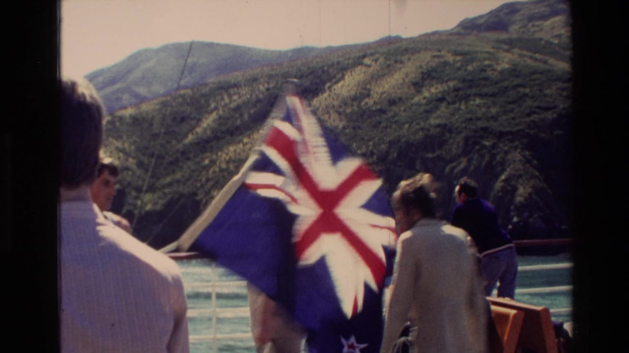 1982 New Zealand home-movie frame showing passengers on a boat deck with a New Zealand flag and steep green mountains beyond the water.