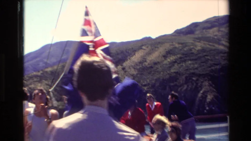 Passengers gather beneath a windblown New Zealand flag during a 1982 boat journey through a mountainous New Zealand waterway.