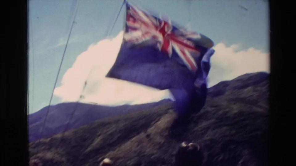A windblown New Zealand flag fills much of the frame in this 1982 home-movie view from a boat, with mountains and sky behind it.