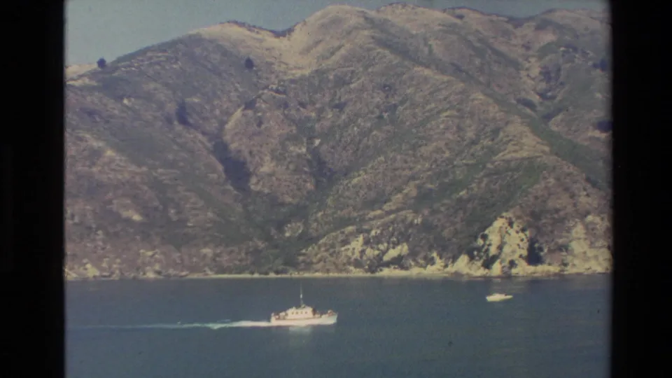 Wide 1982 New Zealand travel-film view of a small vessel moving across blue water beneath steep, sunlit mountain slopes.