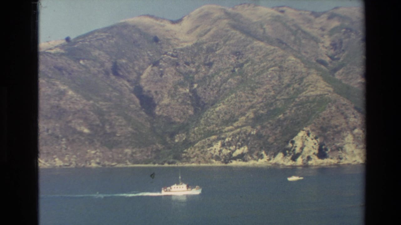 1982 New Zealand 8mm frame showing a small white boat moving across blue lake water below rugged mountain slopes.
