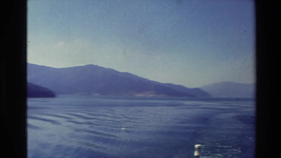 Wide 1982 New Zealand lake view from a moving vantage, with distant mountains and rippling wake across the water.