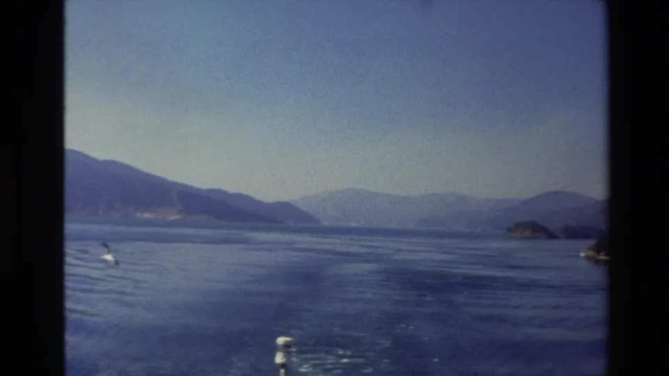 Vintage 8mm New Zealand frame of a broad blue lake, distant hills, small islands, and wake marks from the moving boat.