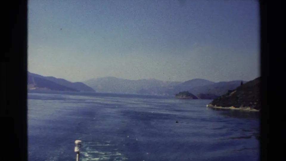 1982 New Zealand 8mm frame looking across a blue mountain lake between sloping shorelines under a pale sky.