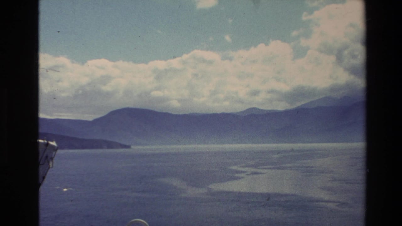 1982 New Zealand 8mm frame showing calm coastal water, hazy mountain ridges, cloud cover, and a small boat edge at left.