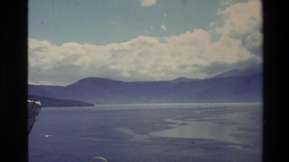 1982 New Zealand 8mm frame showing calm coastal water, hazy mountain ridges, cloud cover, and a small boat edge at left.