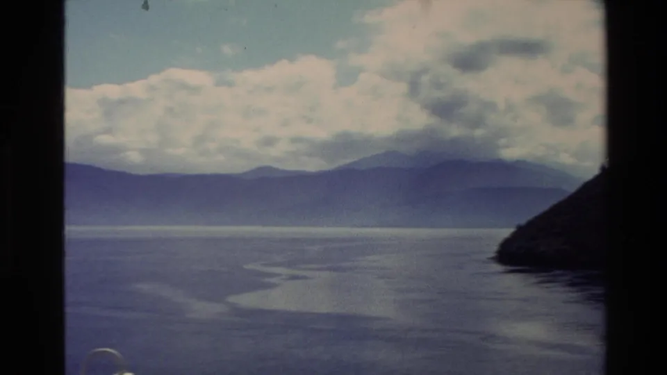 1982 New Zealand 8mm frame with broad water, distant mountains, layered clouds, and a dark vertical film-strip divider through the scan.