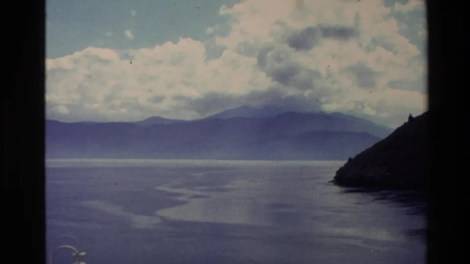 1982 New Zealand 8mm frame showing calm water, hazy mountain silhouettes, clouds, and a dark coastal hillside entering from the right.